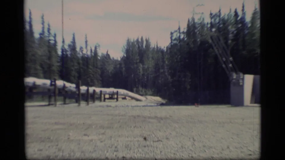 1977 Alaska 8mm frame showing a gravel clearing, timber-supported pipeline, and dark evergreen forest beyond.