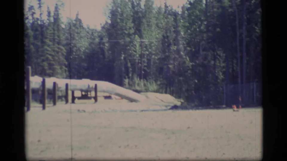 1977 Alaska home-movie frame with a low pipeline crossing cleared ground in front of dense conifer trees.