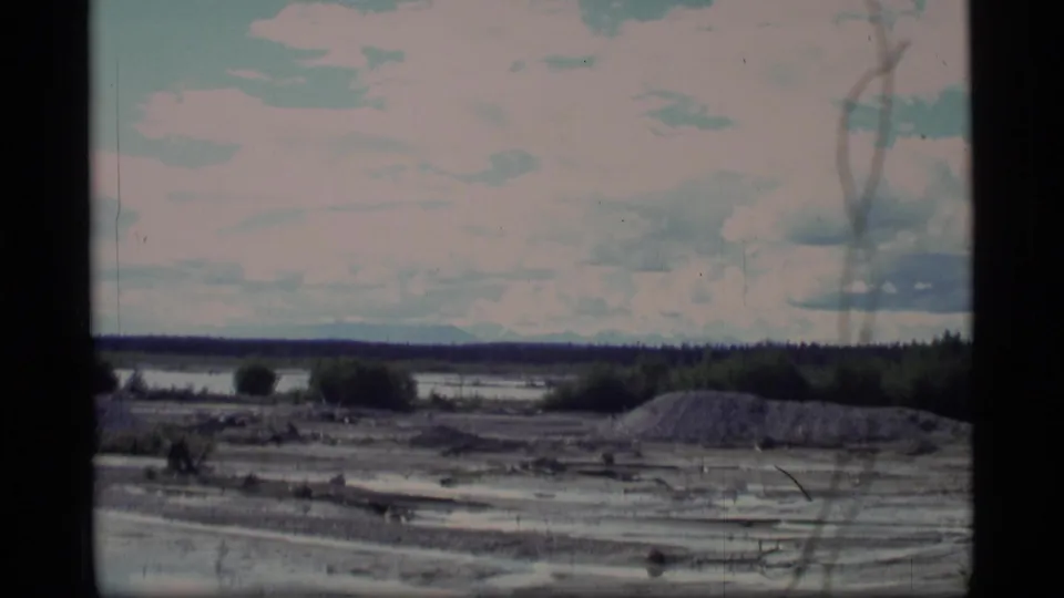1977 Alaska 8mm frame showing muddy water, low brush, gravel piles, distant forest, and cloudy sky.