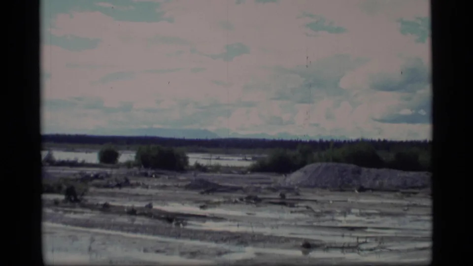 1977 Alaska home-movie frame looking across a waterlogged clearing toward brush, gravel mounds, and a distant forest line.