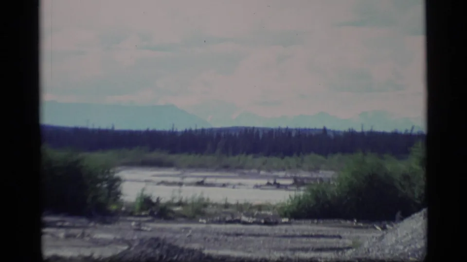 1977 Alaska travel frame with gravelly river flat, brush, a dark tree line, and hazy mountains under overcast light.