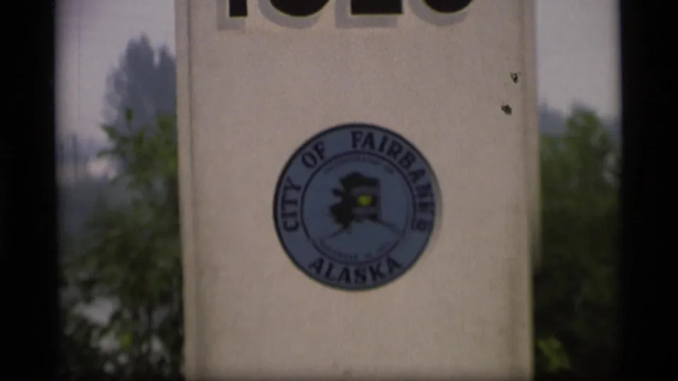 White Alaska Highway marker with the City of Fairbanks Alaska seal and partial top lettering, with trees and sky behind it.