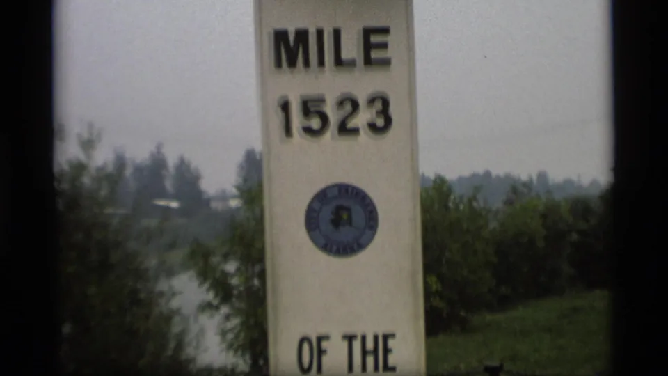 1977 Alaska Highway marker showing Mile 1523 above a City of Fairbanks seal, with river, shrubs and distant trees in the background.