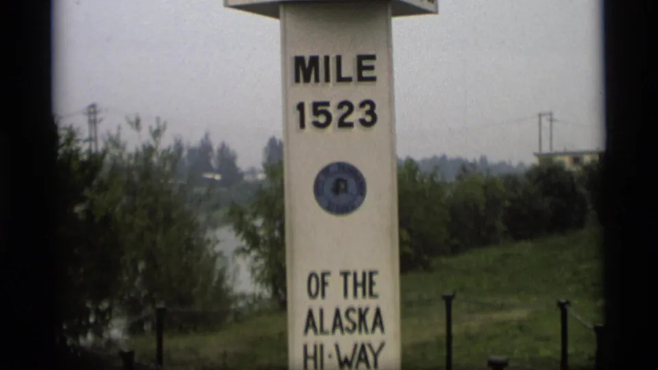 Wider 1977 8mm view of the Mile 1523 Alaska Highway marker, including the Fairbanks seal, lower highway text, grass and distant utility poles.