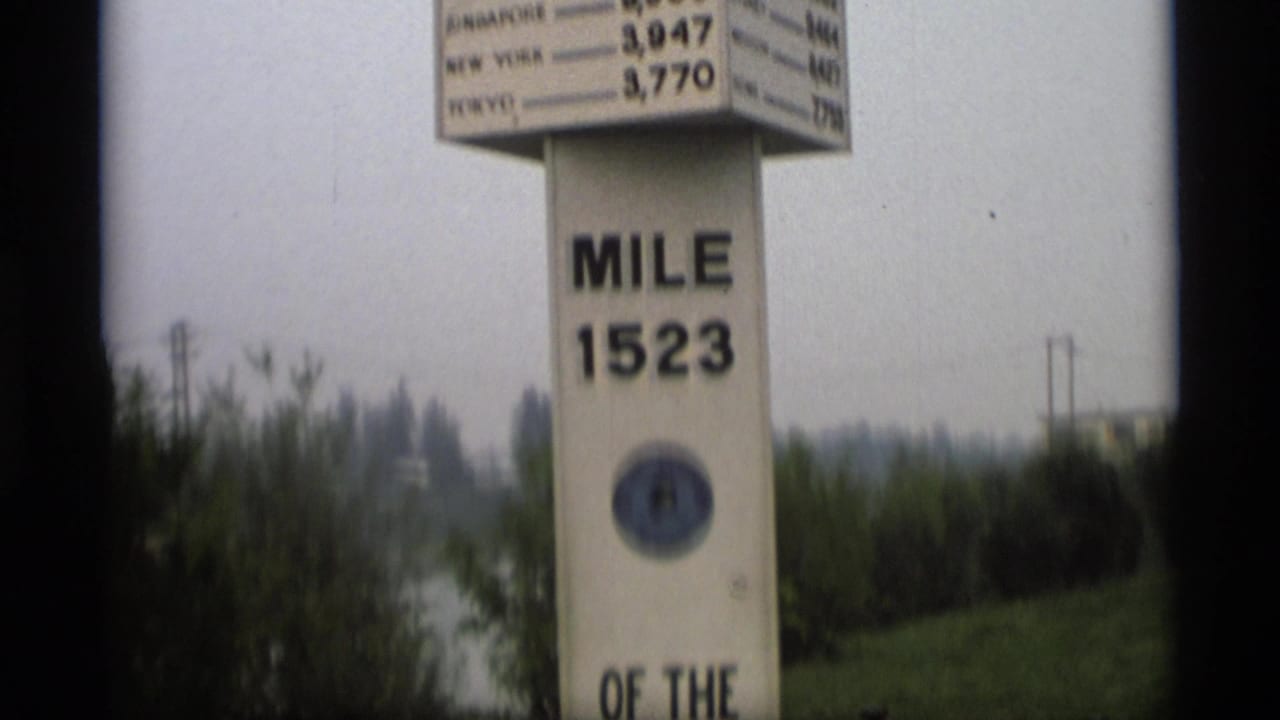1977 Alaska home-movie frame showing a tall mile 1523 marker with a distance sign, trees, and gray sky.