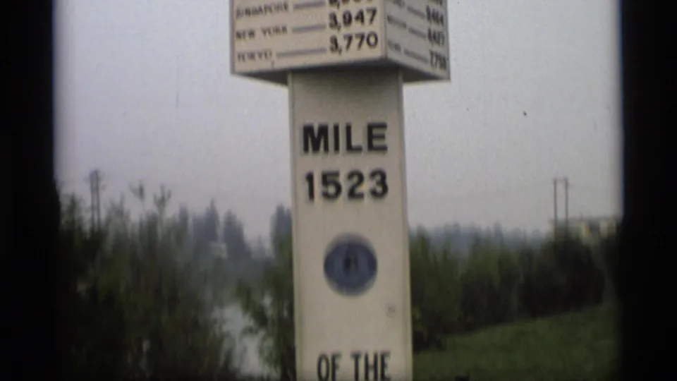 1977 Alaska home-movie frame showing a tall mile 1523 marker with a distance sign, trees, and gray sky.