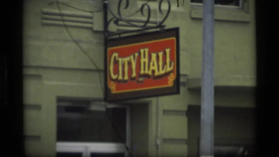 1977 Alaska home-movie frame showing a City Hall hanging sign on a greenish building exterior.