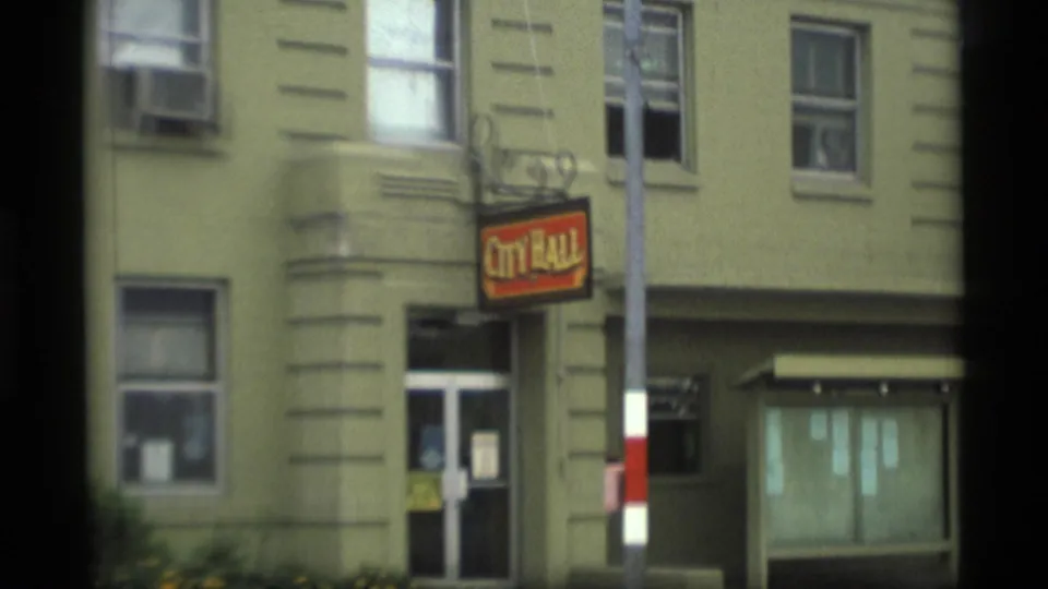1977 Alaska frame showing a small City Hall exterior with a hanging sign, windows, entrance, and roadside pole.