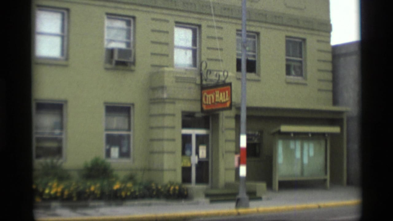 Green two-story building with a City Hall sign, sidewalk, flower bed, and utility pole in front.