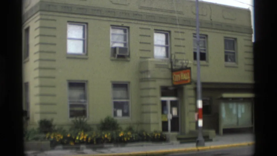 1977 Alaska street view of a city hall facade with sign, covered entrance, windows, planted flowers, and a foreground pole.
