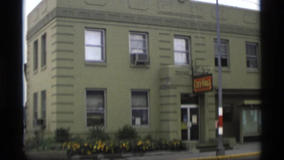 1977 Alaska home-movie frame with an angled view of the city hall building, sign, windows, entrance area, flowers, and curb.