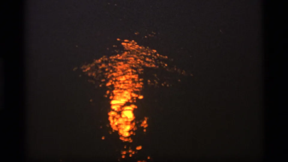 Dark 1977 Alaska home-movie frame showing orange sunset light reflected across rippling water, likely on a lake.