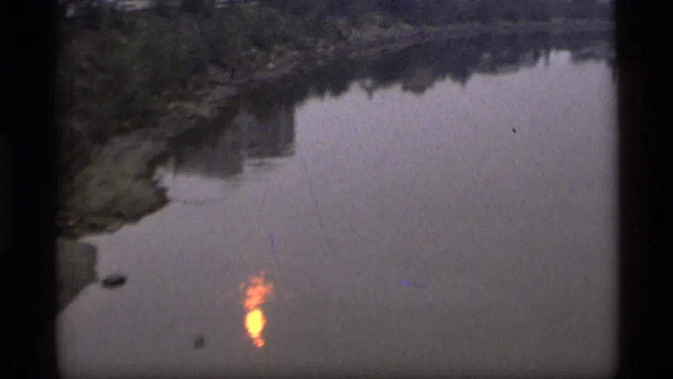 Alaska river water with a small orange sun reflection near a dark, tree-lined shore.