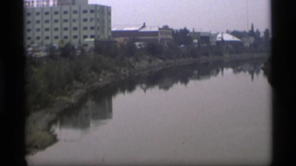 1977 Alaska home-movie view of a calm river, tree-lined bank, and distant low buildings.
