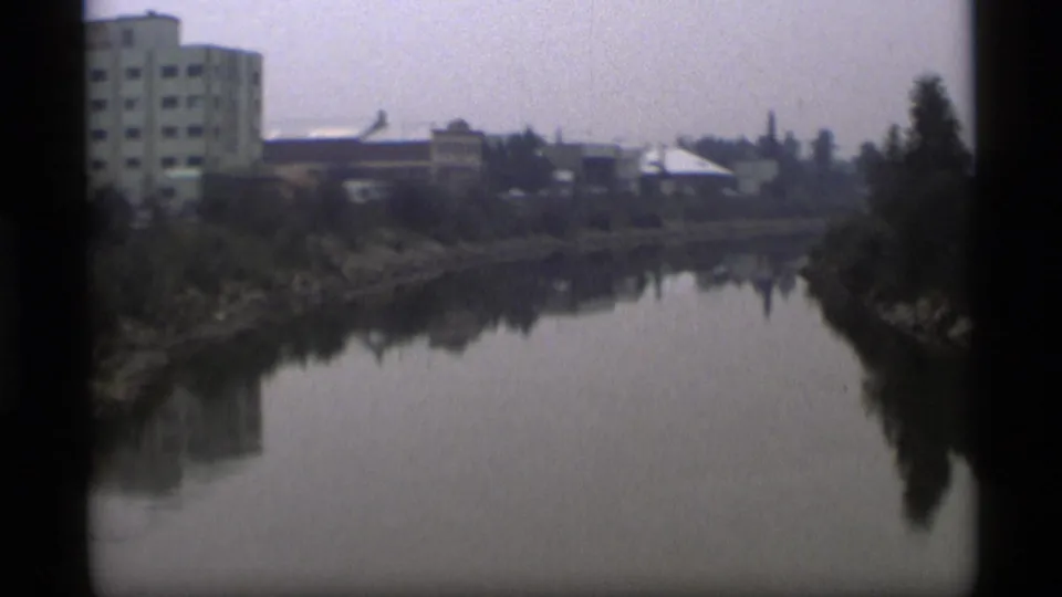 Wide 8mm frame of an Alaska riverside landscape with trees, calm water, and distant buildings.