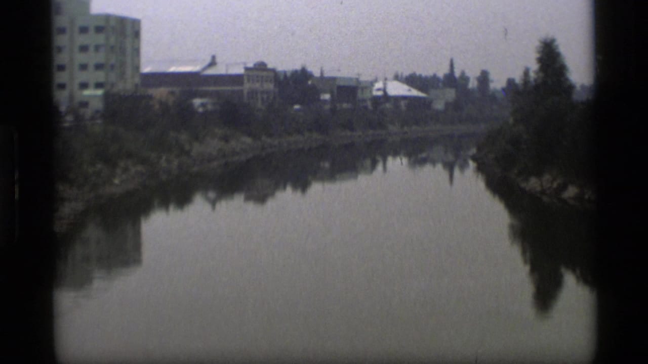 1977 Alaska home-movie frame showing a calm river, shoreline trees, and low urban buildings in soft 8mm focus.