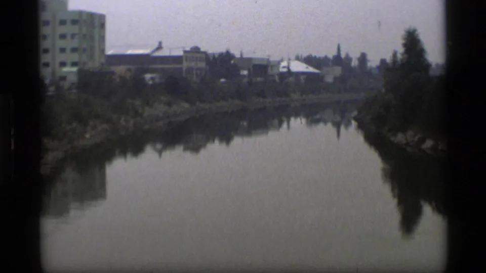 1977 Alaska home-movie frame showing a calm river, shoreline trees, and low urban buildings in soft 8mm focus.
