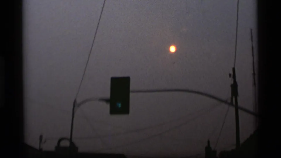 Hazy 8mm view of a bright sun or moon above a suspended traffic signal and utility wires in Alaska, 1977.