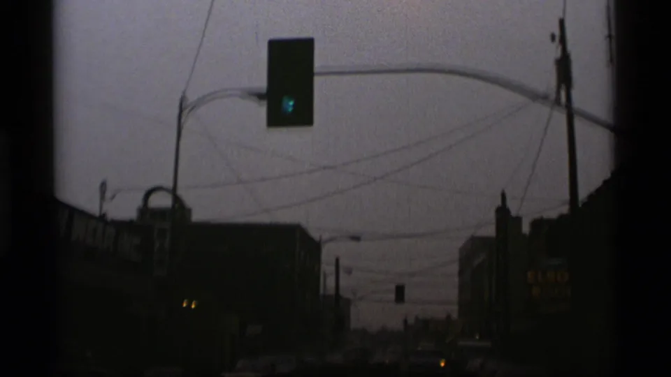 1977 Alaska street scene with a traffic signal, commercial storefronts, overhead wires, and classic cars in dim 8mm exposure.