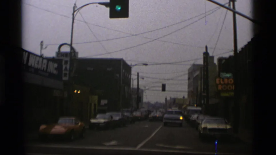 Street-level 1977 Alaska frame showing classic cars, storefront signage, traffic lights, and overhead wires along a commercial road.