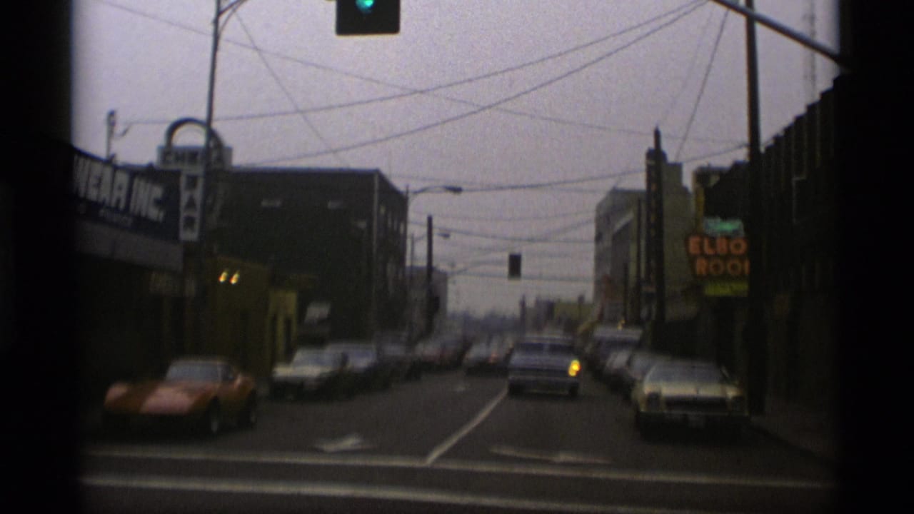 Urban street seen from a vehicle with classic cars, traffic light, power lines, and storefront signage.