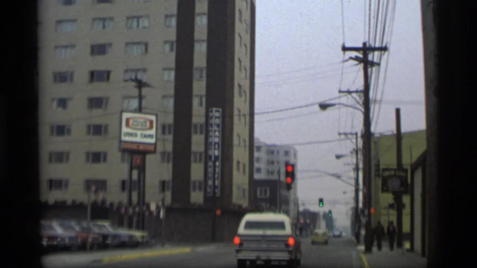 1977 Alaska street view with a tall roadside building, visible used-car signage, parked cars, traffic lights, and distant pedestrians.
