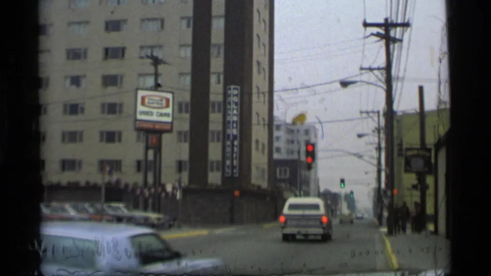 Moving 8mm view of a 1977 Alaska street with a white station wagon, used-car signage, tall building, traffic lights, and pedestrians.