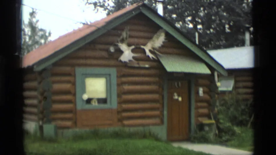 1977 Alaska home-movie frame showing a small log-cabin-style storefront with green trim, antler display, grass, trees, and a walkway.