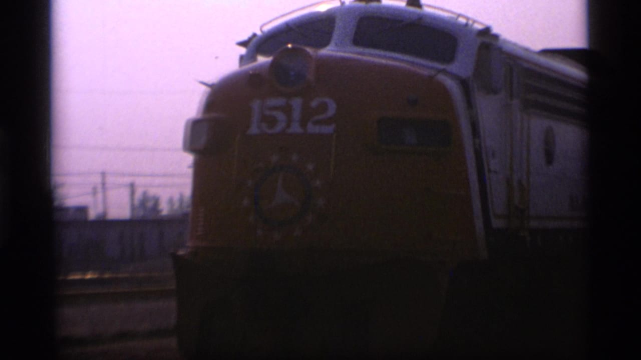 A yellow locomotive marked 1512 appears near the camera in 1977 Alaska home-movie footage, with hazy sky and rail-side structures behind it.