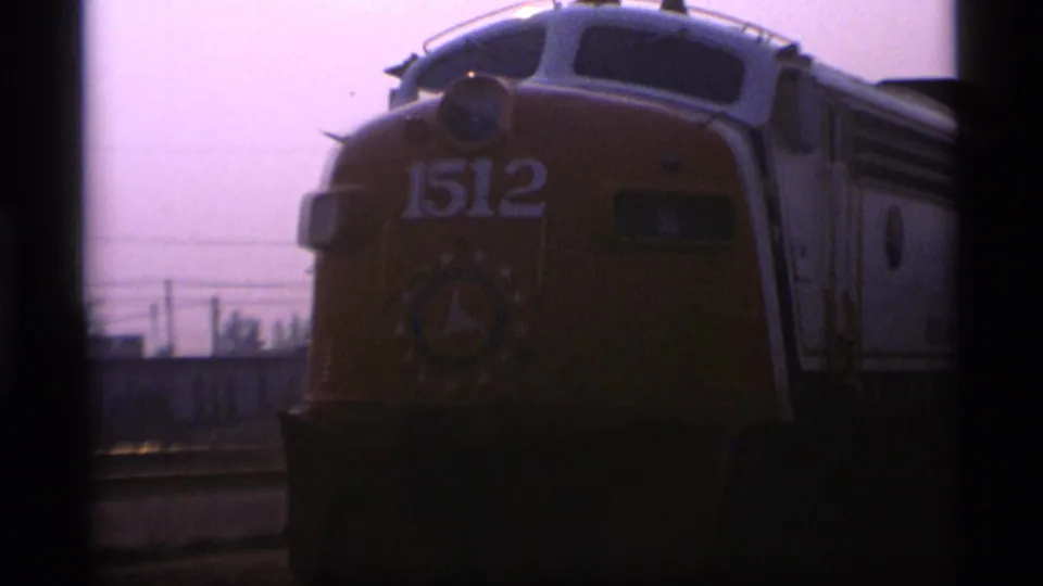 A yellow locomotive marked 1512 appears near the camera in 1977 Alaska home-movie footage, with hazy sky and rail-side structures behind it.
