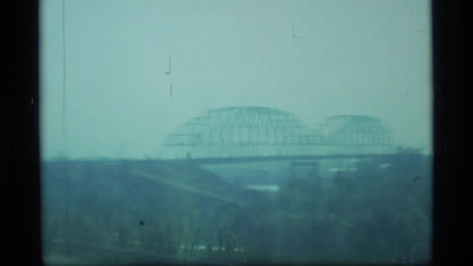 A distant truss bridge spans the landscape in a bluish 1977 Alaska home-movie frame, with low horizon and visible film scratches.