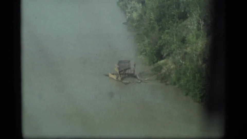 1977 Alaska 8mm frame showing greenish river water, a tree-lined bank, and a small wooden raft-like object near shore.