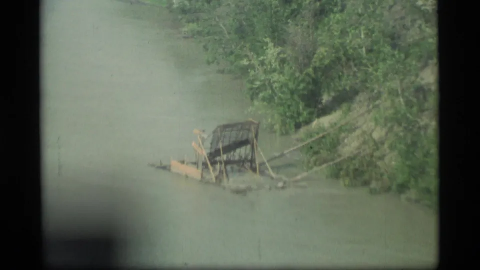 1977 Alaska 8mm frame with a closer view of a small wooden raft-like structure or debris at the edge of a murky river.