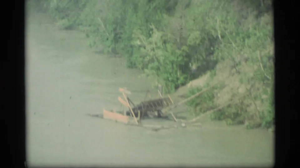 1977 Alaska 8mm frame showing murky river water, a green bank, and a low wooden object partly near the shoreline.