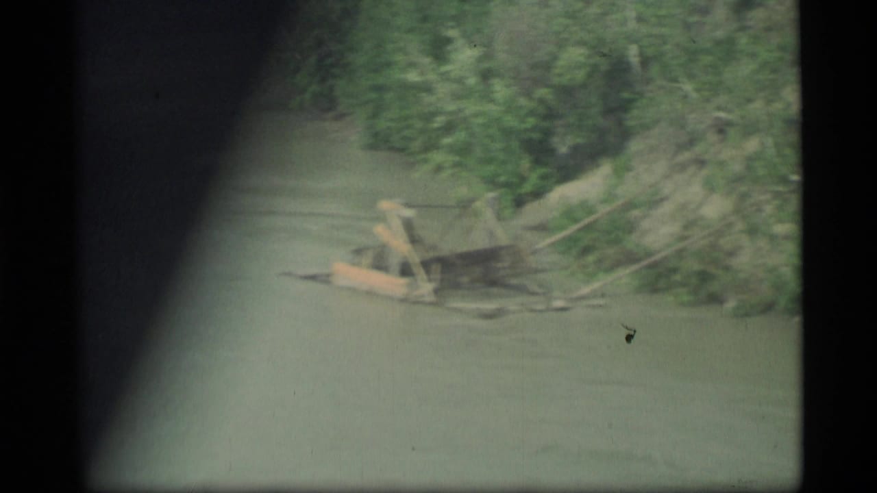 1977 Alaska 8mm view from a moving vehicle near a bridge, looking over water toward a wooded bank and exposed shoreline.