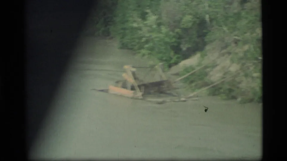 1977 Alaska 8mm view from a moving vehicle near a bridge, looking over water toward a wooded bank and exposed shoreline.
