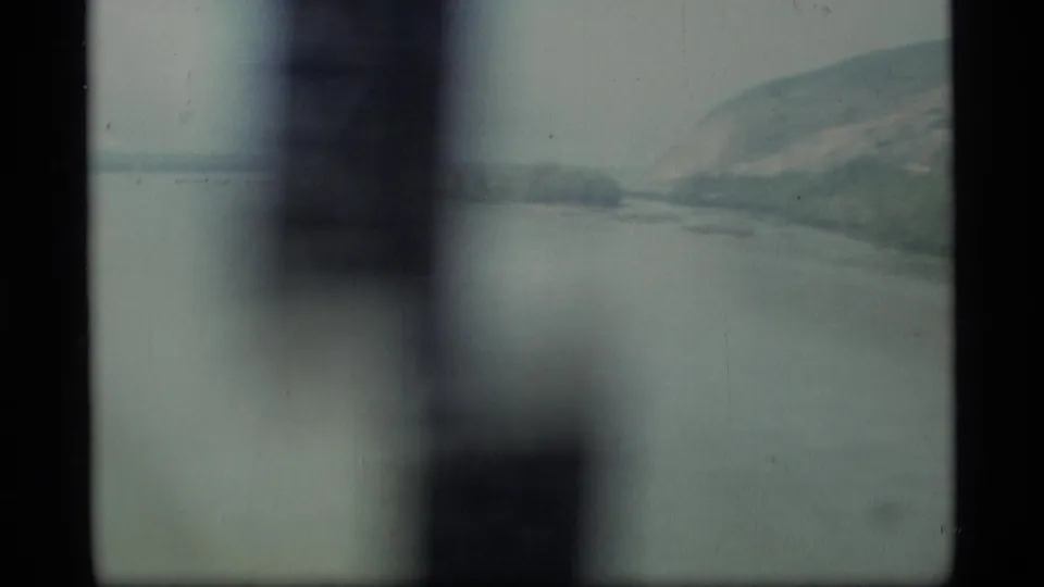 1977 Alaska travel footage looking through dark vertical frame bars across a broad waterway toward hazy hills.