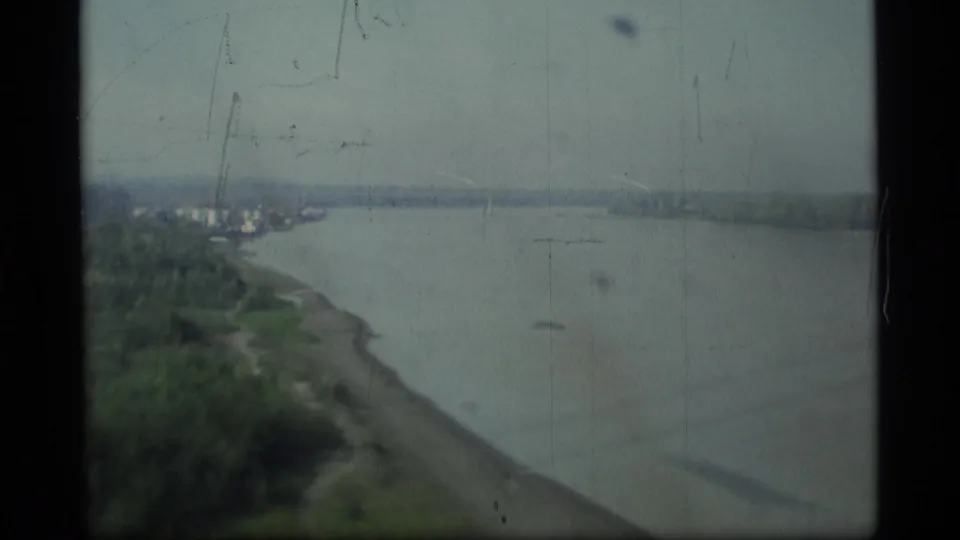 1977 Alaska home-movie view toward a small shoreline settlement, broad water, and faint bridge elements from a moving crossing.