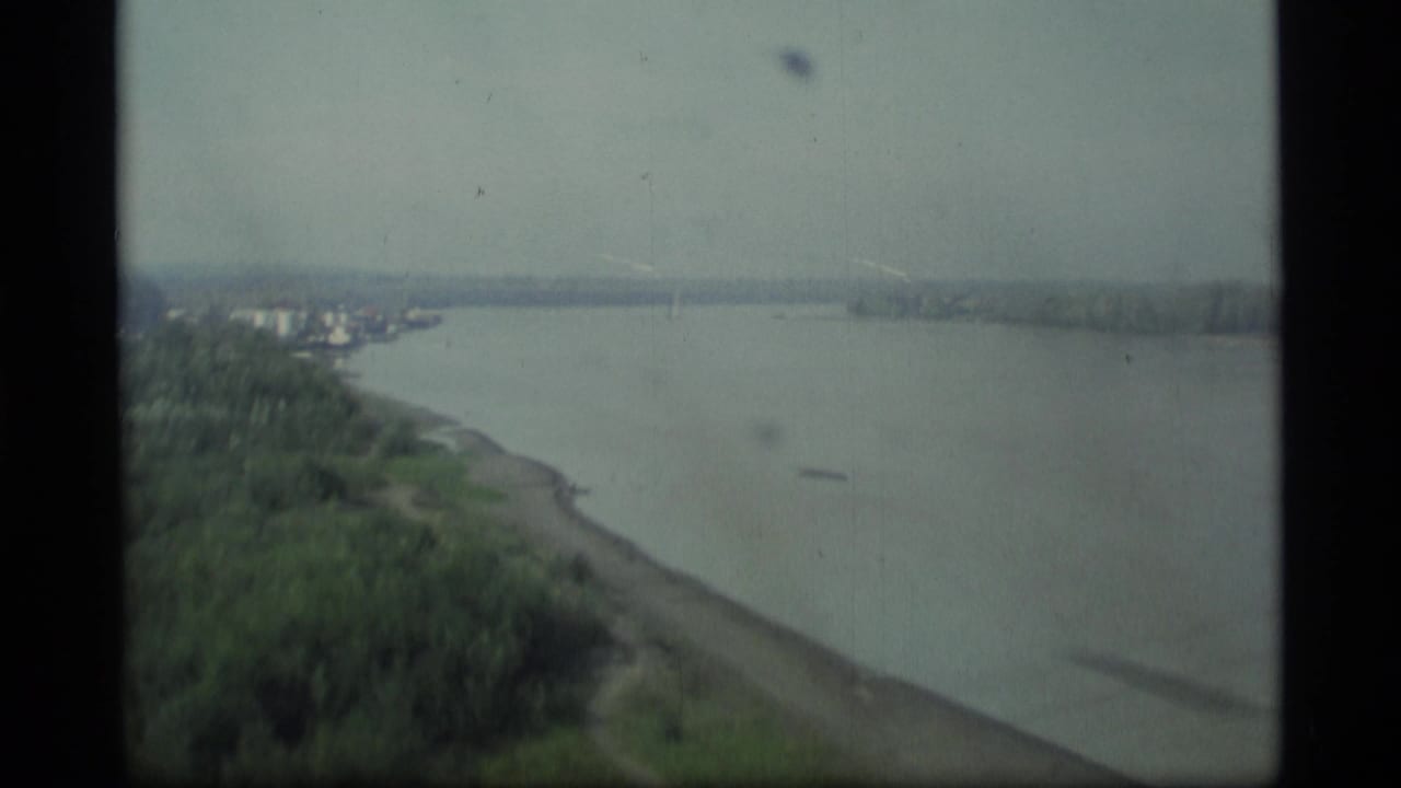 1977 Alaska 8mm frame showing a wide river, green shoreline, distant trees, and a gray stormy sky from a moving viewpoint.