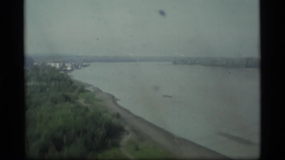 1977 Alaska 8mm frame showing a wide river, green shoreline, distant trees, and a gray stormy sky from a moving viewpoint.