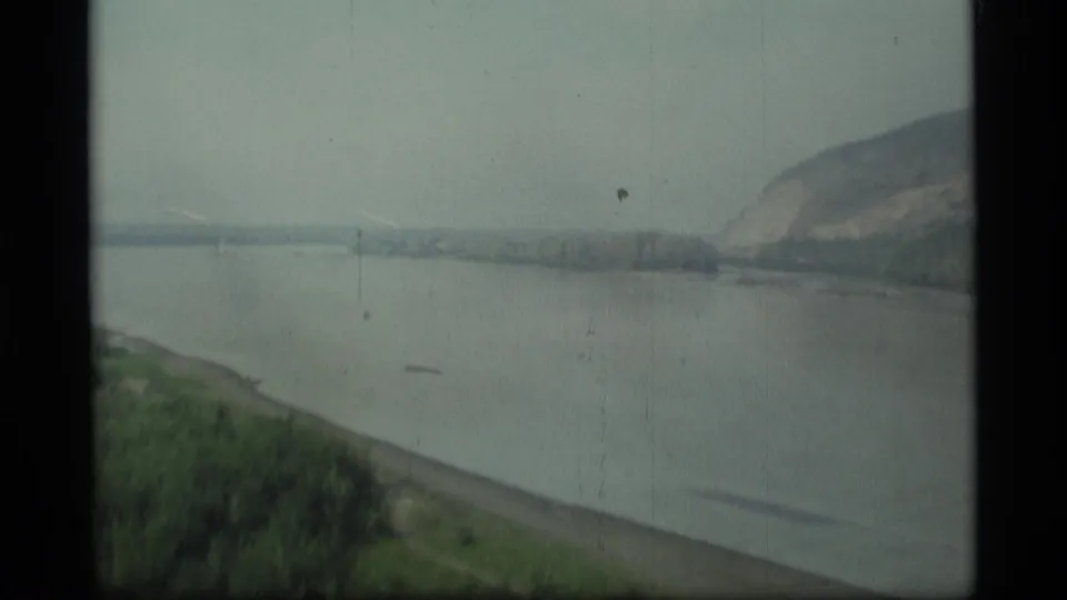1977 Alaska 8mm frame looking across a broad river toward a sloped bank, distant trees, and a low hillside beneath storm clouds.