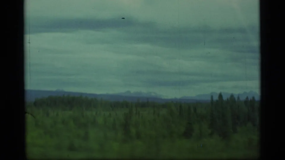 1977 Alaska 8mm frame showing dark green forest, distant low hills, and layered gray storm clouds from a moving travel perspective.