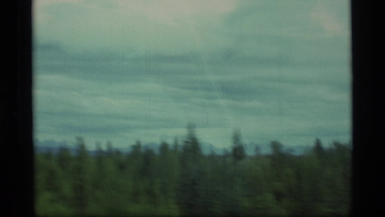 1977 Alaska 8mm travel frame showing blurred green fields, tree line and distant hills through a vehicle window.