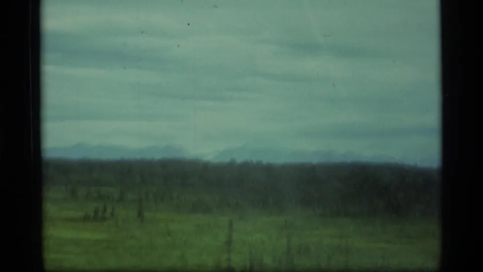 1977 Alaska home-movie frame with open green terrain, wooded horizon and faint mountains beyond a vehicle window.