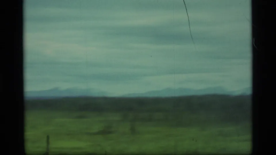 Soft 8mm frame of Alaska countryside in 1977, with blurred grassland, low hills and cloudy sky seen from transit.