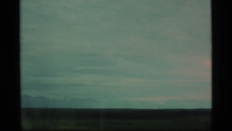 1977 Alaska home-movie frame with a broad cloudy sky, dark horizon, and faint distant mountain forms.