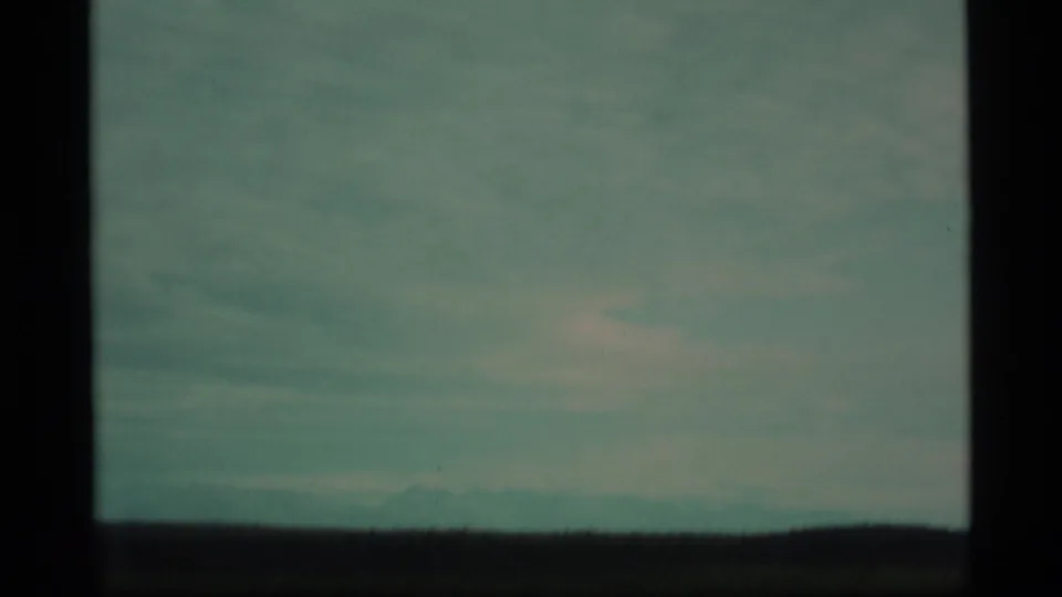 1977 Alaska 8mm frame showing flat green land, a low dark ridge, and pale mountains under hazy cloud cover.