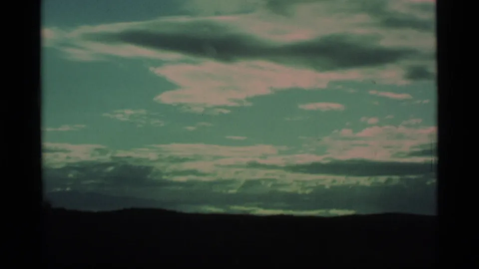 1977 Alaska home-movie frame with a dark ridge, layered cloud banks, and blue-green sky from a moving landscape view.