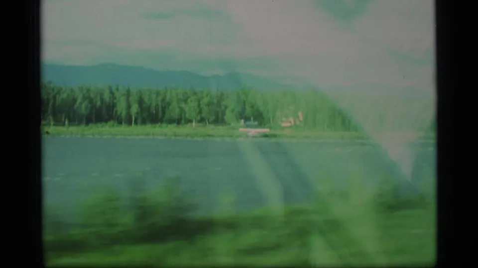 1977 Alaska travel frame showing water, dense forest, distant mountains, and blurred foreground from a moving vehicle.