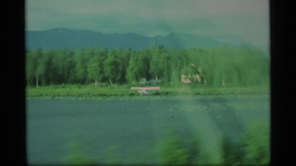 1977 Alaska 8mm frame showing a lake, evergreen shoreline, and blue mountain ridge, softened by motion from a moving viewpoint.